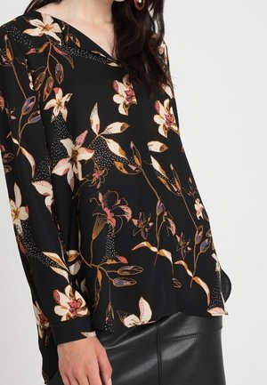 Sort blomstret bluse med store hvide og pink blomster med brune detaljer, lange ærmer og v-halsdesign, parret med en sort nederdel.