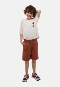 T-shirt beige clair avec un motif de palmier noir, associé à un short couleur rouille avec une taille élastique et une grande poche latérale. Baskets multicolores.