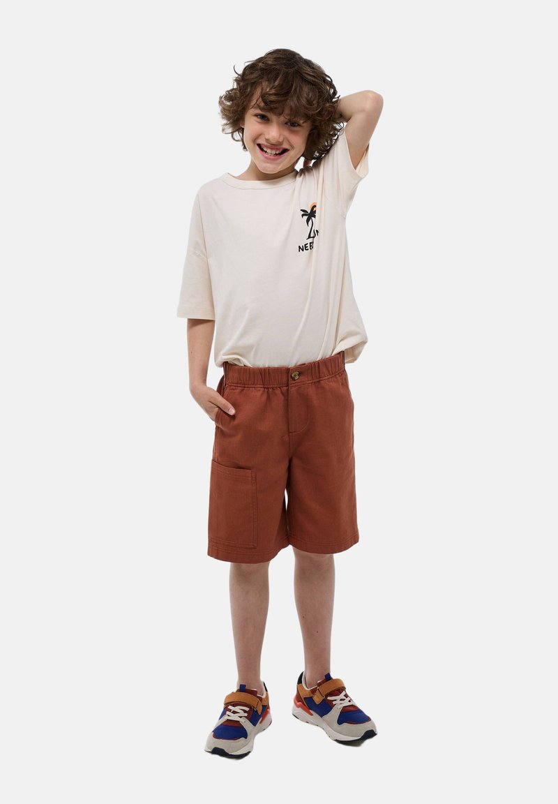 T-shirt beige clair avec un motif de palmier noir, associé à un short couleur rouille avec une taille élastique et une grande poche latérale. Baskets multicolores.