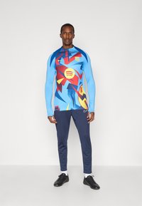 Niebieska bluza sportowa z długim rękawem, z abstrakcyjnym wzorem w kolorach czerwonym, żółtym i niebieskim, w zestawie z granatowymi spodniami i czarnymi butami.