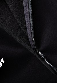 Giacca nera con zip dotata di un morbido rivestimento interno, tirazip testurizzato e logo stampato. I dettagli in primo piano mettono in evidenza cuciture e materiali.