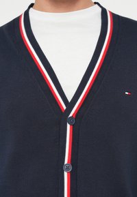 Ναυτικό κασκόλ με κόκκινες και λευκές ρίγες στην λαιμόκοψη, διαθέτοντας σχέδιο σε V, κλείσιμο με κουμπιά και λεπτομέρεια λογότυπου στο αριστερό στήθος.