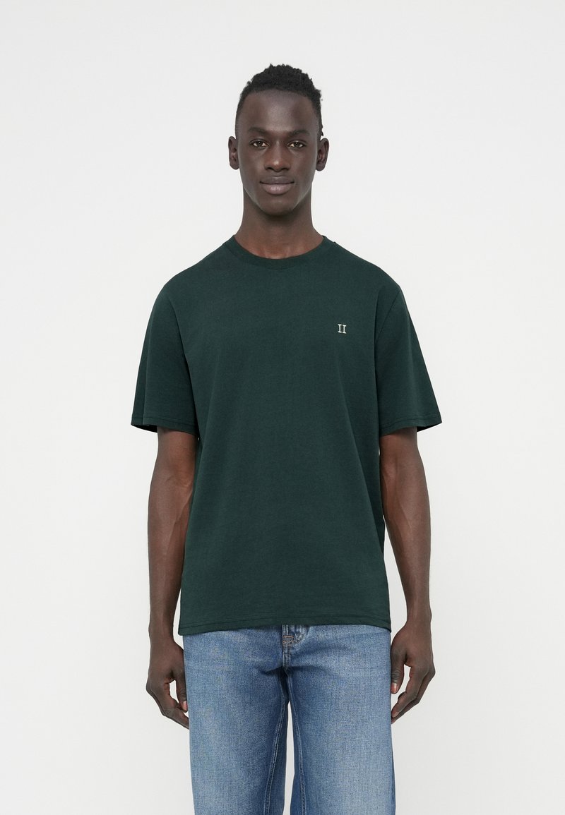 T-shirt en coton vert foncé à manches courtes, col rond, avec un petit logo blanc "II" sur le côté gauche de la poitrine. Porté avec un jean bleu clair.