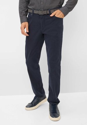 Marineblaue Chinos mit schmaler Passform aus Baumwollmischgewebe; ausgestattet mit fünf Taschen und Gürtelschlaufen; kombiniert mit dunklen Sneakers.