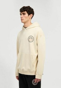 Sudadera color crema con un bolsillo frontal y un logo circular en el pecho. Tejido suave, mangas largas y capucha con cordón.