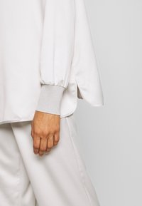 Mano descansando al lado, vistiendo pantalones blancos holgados y una camiseta blanca de manga larga con puños acanalados y detalle de abertura lateral.