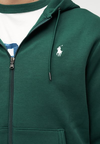 Άνδρας που φοράει σκούρο πράσινο φούτερ με φερμουάρ και λογότυπο αλόγου πόλο με λευκή κεντημένη λεπτομέρεια και λευκό T-shirt από μέσα.