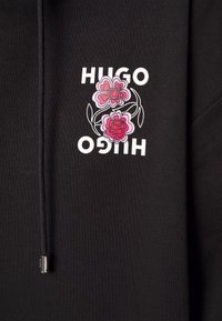 Černá mikina s kapucí se bílým textem "HUGO" zrcadleným vertikálně a dvěma stylizovanými květinami v růžové a červené barvě se zeleně obrysem stonků mezi textem.