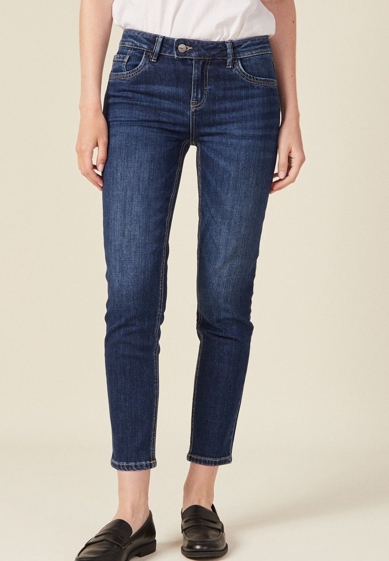 Tmavě modré džíny v úzkém střihu, s středně vysokým pasem, pěti kapsami a nohavicemi s manžetami. Mají ozdobné švy a kovové zapínání na knoflík.