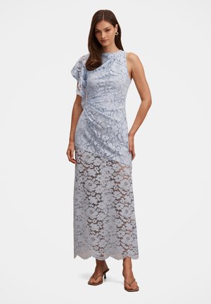 Žena u svijetloplavoj maksi haljini od čipke s jednim naboranim rukavom i prozirnim cvjetnim uzorkom, uparena s bež sandalama na petu.