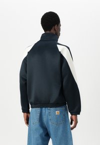 Casaco azul-marinho com detalhes brancos, gola alta e cintura elástica. A vista traseira revela uma textura suave e painéis contrastantes. Jeans de denim são visíveis.