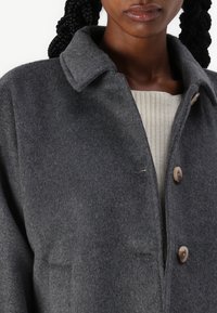 Manteau en mélange de laine gris foncé, avec un col, deux poches avant et une fermeture à boutons. Tissu texturé avec une finition lisse.