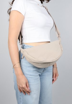 Donna che indossa jeans di un blu chiaro e una maglietta cropped bianca, con una borsa a tracolla beige appoggiata all'anca su uno sfondo semplice.