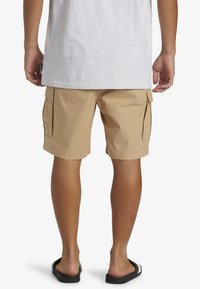 Shorts cargo beiges avec poches latérales, fabriqués en tissu léger, présentant une surface texturée et une coupe décontractée. Portés avec des sandales noires.