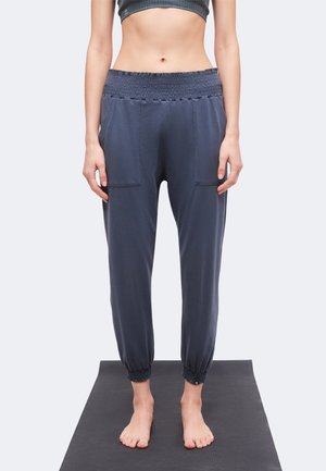 Persona de pie descalza sobre una esterilla de yoga, con pantalones jogger azul marino de talle alto con cintura y puños elásticos, visible desde el pecho hacia abajo.