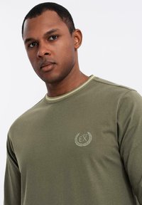 Olivengrünes Langarmshirt aus Baumwolle, mit rundem Halsausschnitt und gesticktem, kreisförmigem Logo auf der linken Brust aus leichtem Faden.