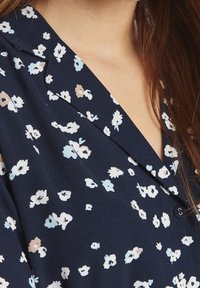 Närbild av en person som bär en marinblå blus med ett vitt och beiget blommönster och öppen krage, med långt brunt hår synligt.