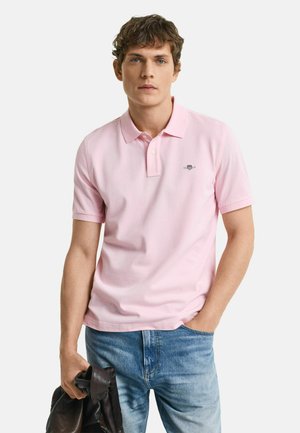 Rosa Polohemd mit Kragen und kurzen Ärmeln, mit einem kleinen gestickten Logo auf der linken Brust. Getragen mit hellblauen Jeans.