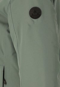 Grön tygjacka med en slät textur, med kontrasterande svarta sömmar och ett cirkulärt svart logopatch på överarmen.
