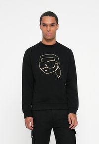 Uomo che indossa una felpa nera con un disegno dorato di un volto con occhiali da sole oversize e una coda di cavallo su uno sfondo bianco.