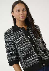 Černobílý kostkovaný cardigan s krátkými rukávy, texturovaná látka, špičatý límec, přední knoflíky a boční kapsy.