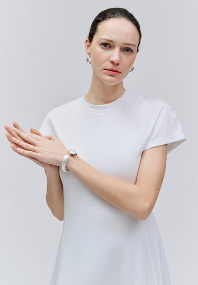 Femme aux cheveux foncés en robe blanche, mains jointes, portant des boucles d'oreilles argentées en anneau et un bracelet argenté épais, sur un fond gris uni.