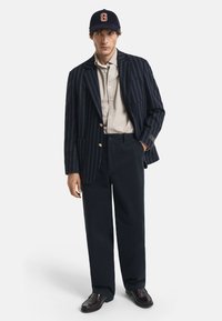 Man die een marineblauwe gestreepte blazer, een beige overhemd met knopen, donkere wijde broek, donkere loafers en een marineblauwe pet met een oranje "G" draagt, staat voor een witte achtergrond.