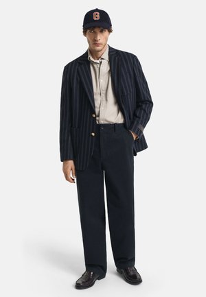 Man i marinblå randig kavaj, beige skjorta med knappar, mörka vida byxor, mörka loafers och marinblå keps med orange "G" som står på vit bakgrund.