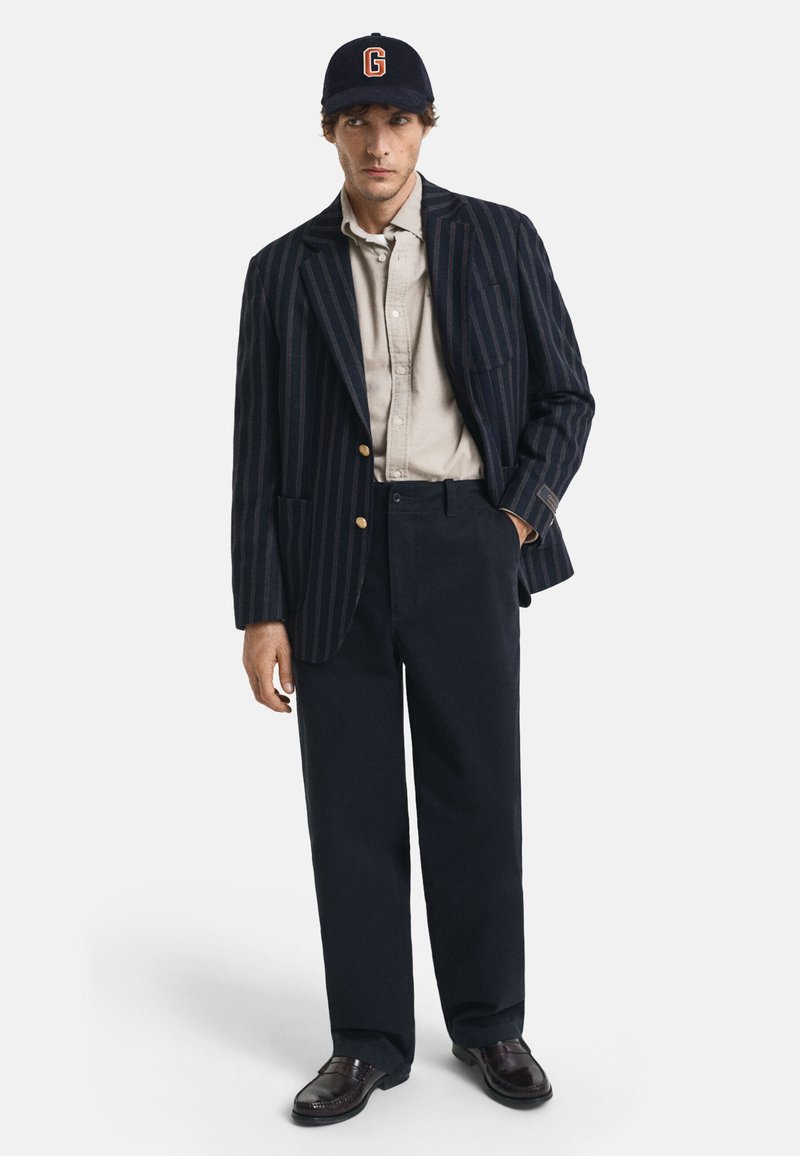 Man die een marineblauwe gestreepte blazer, een beige overhemd met knopen, donkere wijde broek, donkere loafers en een marineblauwe pet met een oranje "G" draagt, staat voor een witte achtergrond.