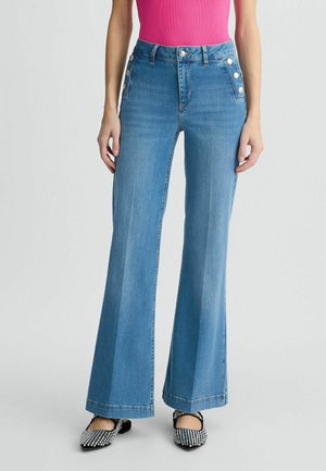 Vrouw draagt lichtblauwe flare jeans met hoge taille en zilveren knopen op de zakken, roze top en zwart-witte geruite platte schoenen.