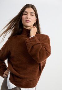 Brązowy sweter dziany z wysokim kołnierzem, ściągaczami przy mankietach i luźnym krojem. Noszony na białej koszuli, z miękkiego, fakturowanego materiału.