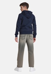 Tiener in een marineblauwe hoodie, grijs vervagende jeans en witte sneakers, staand met de rug naar de kijker, met de handen ontspannen langs de zij.