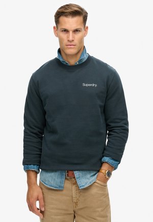 Sudadera de cuello redondo azul oscuro con cuello y puños de canalé, que presenta un pequeño logotipo gris de "Superdry". Llevada sobre una camisa de mezclilla azul claro.