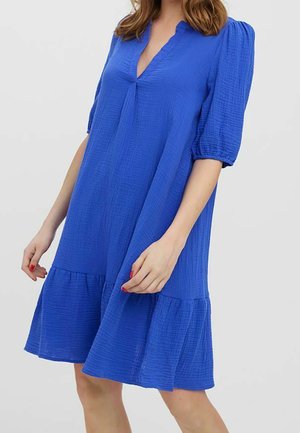 Mujer con un vestido azul de corte suelto, con mangas abullonadas, escote en V y un dobladillo con volantes, de pie frente a un fondo liso.