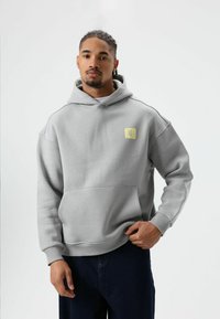 Grijze capuchontrui gemaakt van zacht materiaal, met een voorzak en een klein geel vierkant logo op de linkerborst. Simpel ontwerp met geribde manchetten.