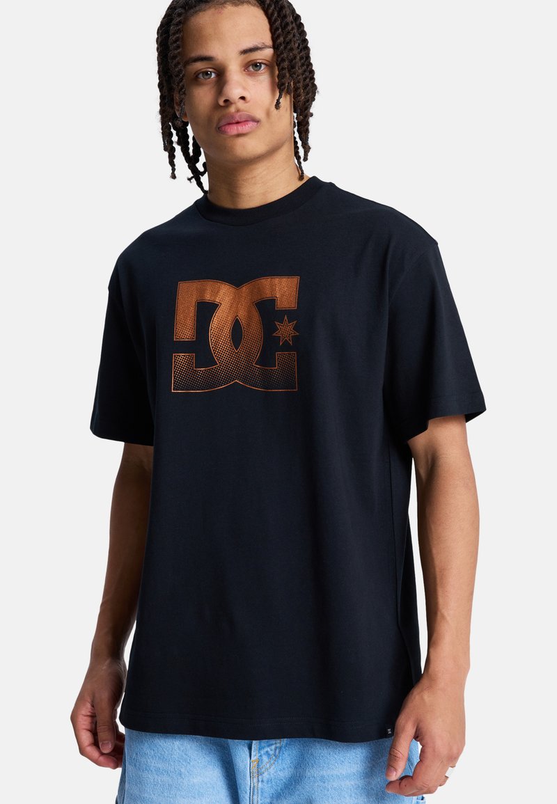 Jeune personne aux cheveux tressés portant un t-shirt noir avec un grand logo DC marron et un jean bleu clair, sur un fond blanc.