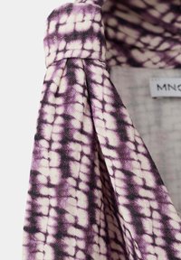 Fialová a bílá látka s vzorem tie-dye přehozená přes háček, zobrazující měkkou texturu a částečnou oděvní etiketu v pozadí.