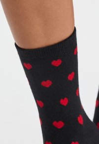 Calze nere con un motivo ripetuto di piccoli cuori rossi. Realizzate in un materiale morbido con un bordo a coste e una texture liscia.