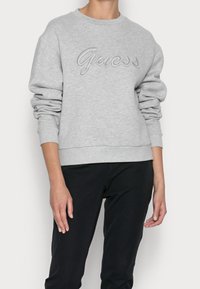 Lichtgrijze sweatshirt gemaakt van zacht materiaal met een ronde hals, voorzien van een verhoogd grijs geborduurd 'Guess'-logo over de borst. Lange mouwen.