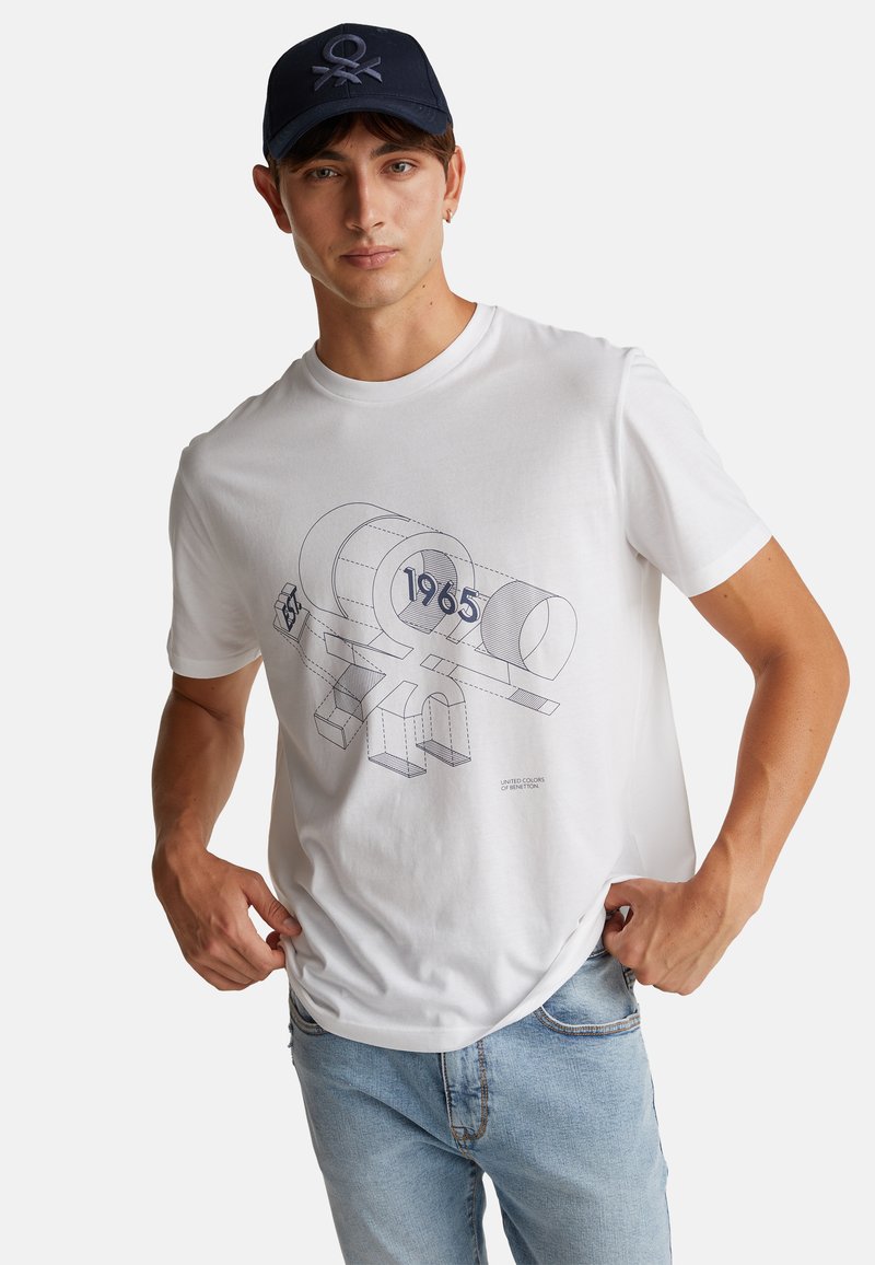 Bílý bavlněný tričko s geometrickou modrou grafikou a textem "1965". Model nosí světle modré džíny a námořnickou čepici s logem.