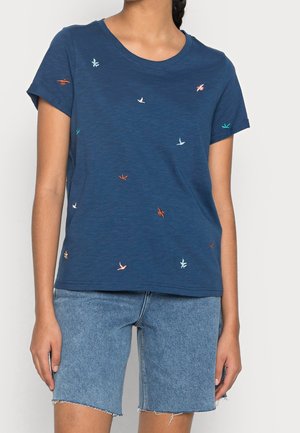 Kobieta ubrana w granatowy t-shirt z małymi wielokolorowymi nadrukami ptaków i jasnoniebieskie dżinsowe szorty z postrzępionymi brzegami.
