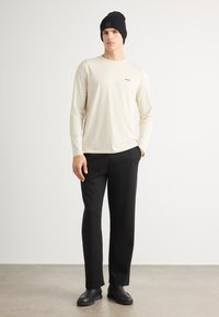 Cămașă bej cu mânecă lungă și logo, fes negru, pantaloni negri și cizme negre; material neted, croială lejeră și design simplu.