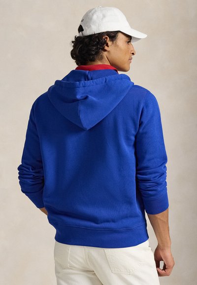 Královsky modrá mikina s kapucí z bavlněné směsi, s klokaní kapsou a žebrovanými manžetami, nošená přes červenou košili, s bílou čepicí.