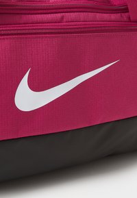 Športová taška Nike s textúrovaným telom vo farbe fuchsie a čiernym dnom, výrazným bielym logom swoosh a dvoma zipsovými priehradkami.