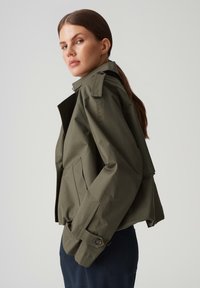 Olijfgroen jasje met een gestructureerd ontwerp, voorzien van een hoge kraag, epauletten, zijzakken en knoopmanchetten. Gladde textuur.