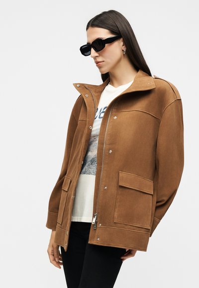 Γυναίκα με μακριά σκούρα μαλλιά, που φοράει μαύρα γυαλιά ηλίου, καφέ ζακέτα με φερμουάρ και μεγάλες τσέπες, λευκό μπλουζάκι με γραφικό σχέδιο και μαύρο παντελόνι.