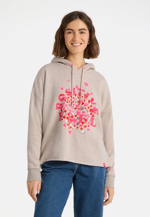 Young woman smiling, wearing beige hoodie with pink and red heart patterns and blue jeans, standing against a plain white background.