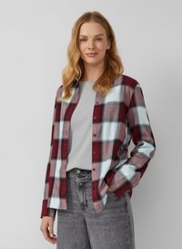 Kobieta z falującymi blond włosami, nosząca bordową i białą koszulę w kratę zapinaną na guziki na lekko szary t-shirt oraz szare dżinsy, uśmiechająca się delikatnie.