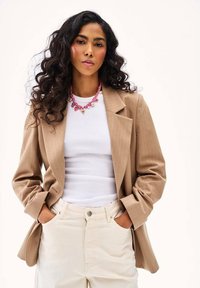 Žena s dlhými kučeravými vlasmi v béžovom blejzri s prúžkami, bielom tričku, krémových nohaviciach a ružovom náhrdelníku srdcia, ruky vreckách.