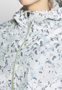 Kevyt takki, jossa on harmaa ja pastellisävyinen abstrakti kuvio, varustettu vetoketjulla ja huppulla. Materiaali näyttää sileältä ja siinä on hieman kiiltoa.